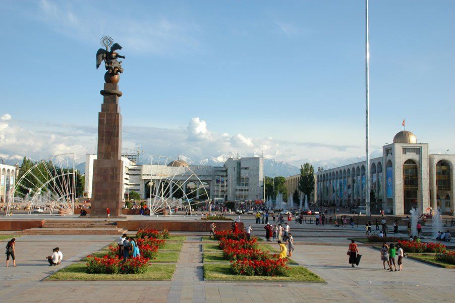 Ala-Too Square in Bishkek, Kyrgyzstan, featuring a monument, fountains, buildings, and people enjoying the sunny day.