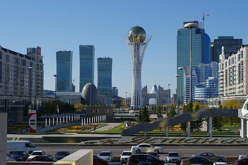 Cityscape of Astana, Kazakhstan, featuring tall modern buildings, with the iconic Bayterek Tower prominent in the center, blue sky.