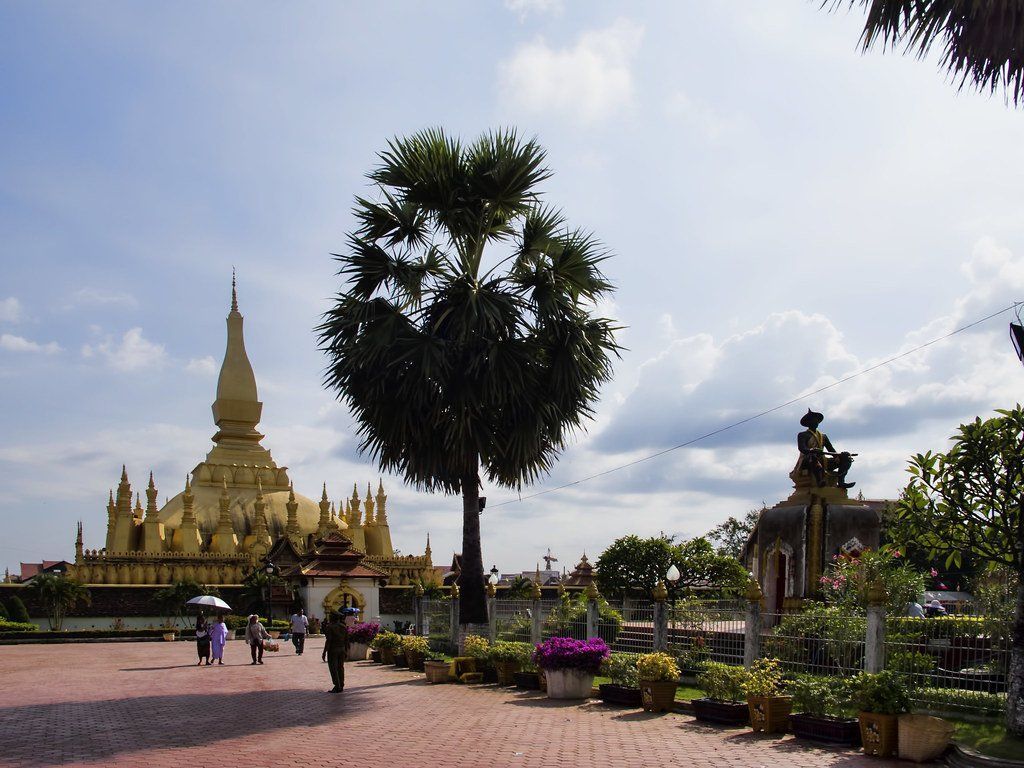 Golden stupa and temple in a park-like setting under a blue sky. Several people stroll by, with a prominent palm tree.