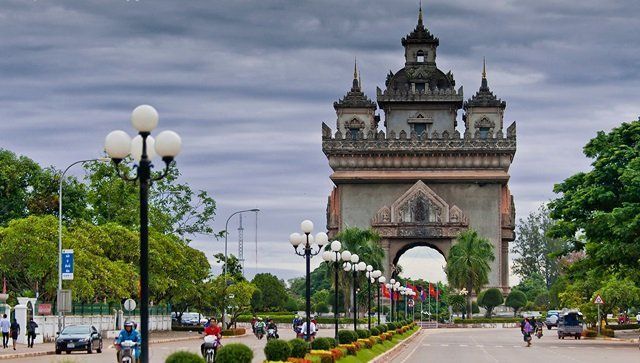 Patuxai monument in Vientiane, Laos, a large arch with ornate details, viewed from a tree-lined avenue on a cloudy day.