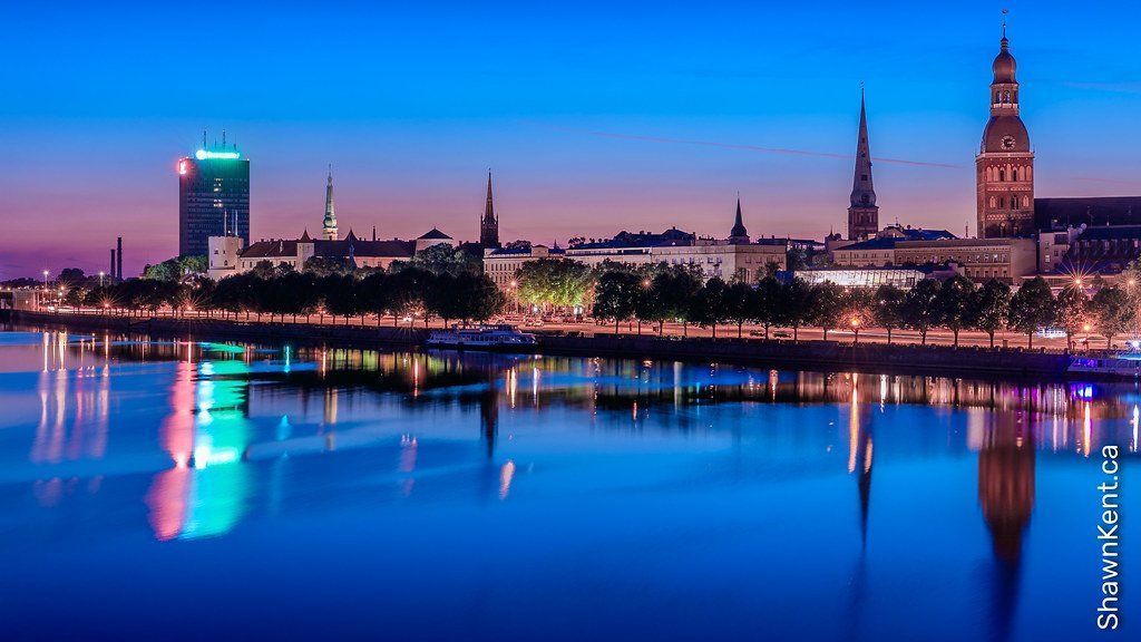Nighttime skyline of Riga, Latvia, reflected in the still water of a river. Buildings of varying heights and architectural styles silhouetted against a dusky blue sky.