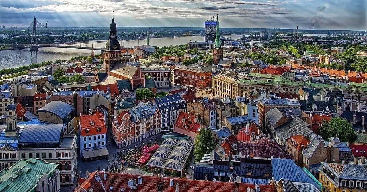 Aerial view of Riga, Latvia, showcasing a cityscape with red-tiled roofs, church spires, a river, and bridges under a partly cloudy sky.