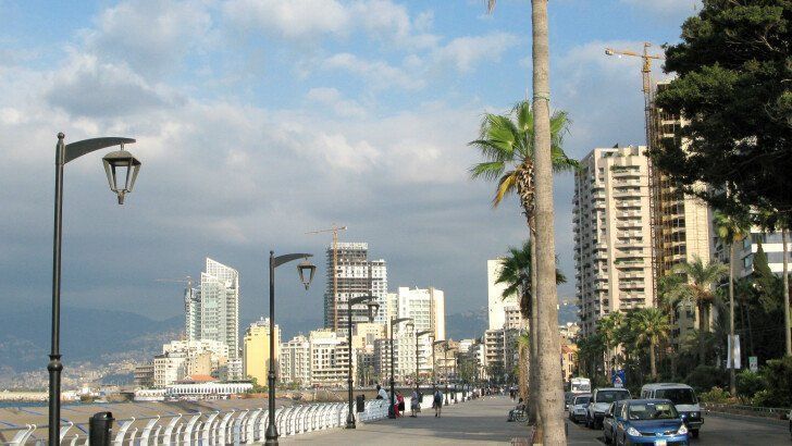 Waterfront view in Beirut, Lebanon, with buildings, palm trees, a seaside promenade, and cars along the road under a cloudy sky.