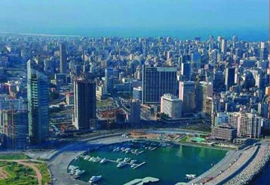 An aerial view of a bustling city skyline, with a marina in the foreground and ocean in the distance under a clear blue sky.