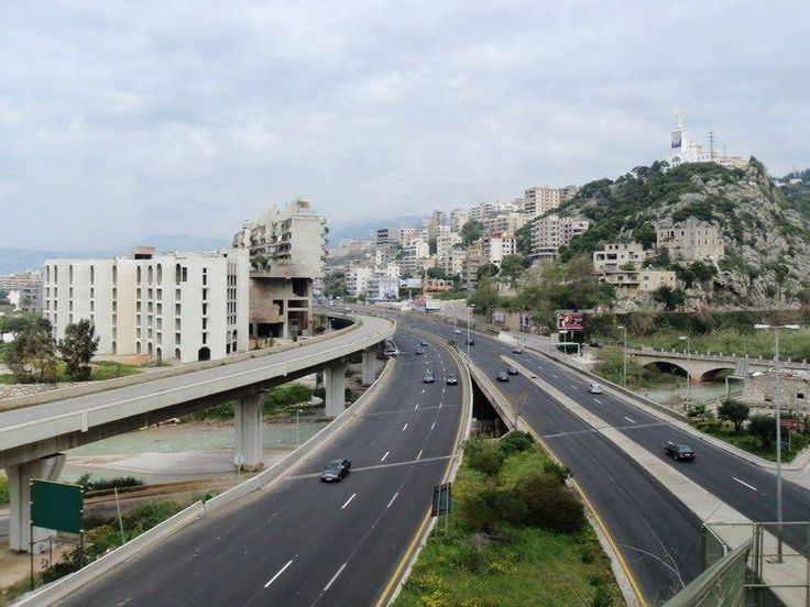 Elevated highway with cars, passing in front of a cityscape of buildings and a hill with a structure on top, under a cloudy sky.
