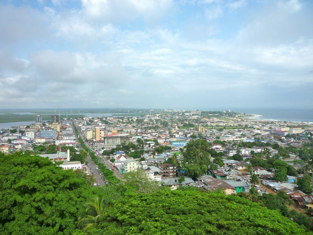 Overhead view of a coastal city with buildings lining a main road, lush green trees in the foreground, and the ocean in the background.