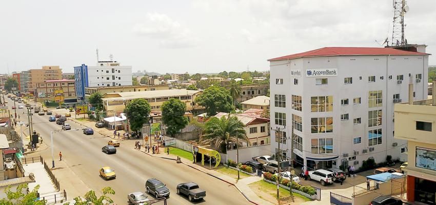 An aerial view of a city street with vehicles, buildings, and trees. A tall white building is on the right side.