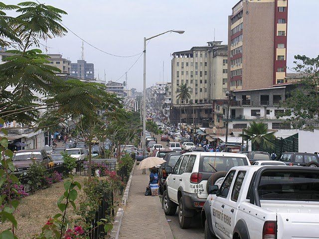 Busy city street in Africa. Cars line a road with tall buildings in the background; a pedestrian walks on the side.