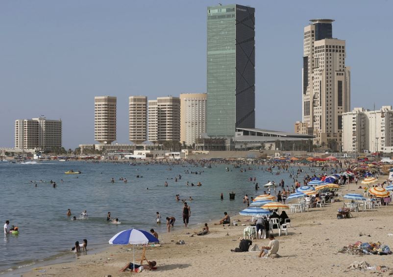 Beach scene in Tripoli, Libya with many people swimming and sunbathing. Skyscrapers line the background on a sunny day.
