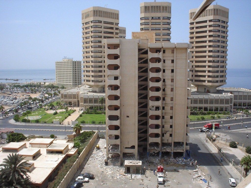 A damaged building in Tripoli, Libya, with several other beige skyscrapers in the background, and a view of the sea.