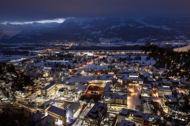 Nighttime view of a town nestled in a valley, illuminated by lights with snowy roofs and mountains in the background under a dark sky.