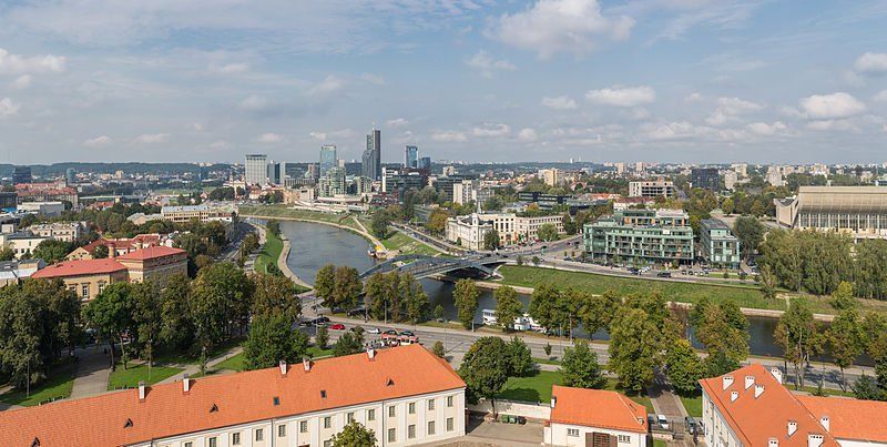 A panoramic view of Vilnius, Lithuania, with a river, buildings, and a skyline under a partly cloudy sky.