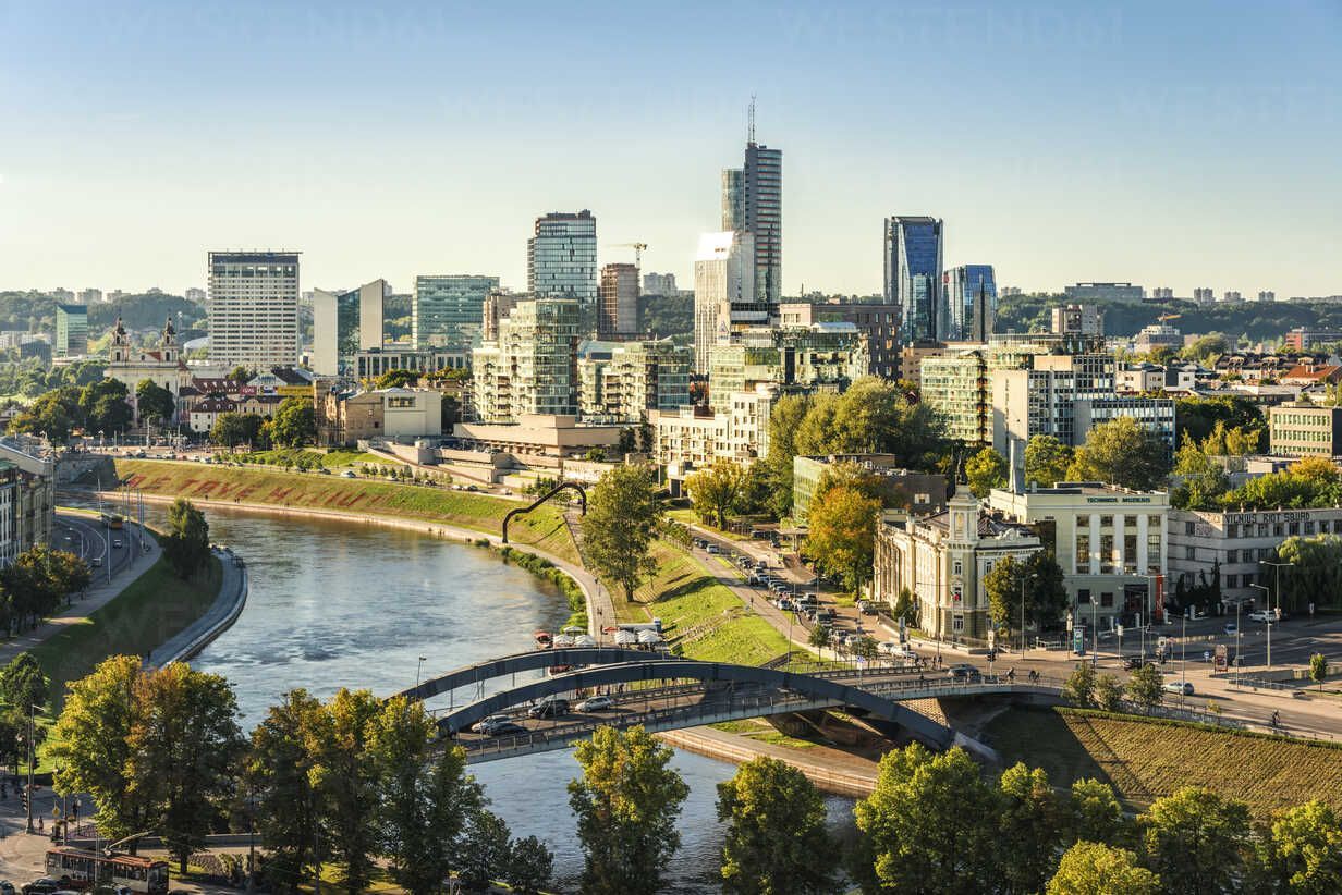 Cityscape of Vilnius, Lithuania, featuring modern skyscrapers and a river with arched bridges, seen under a bright blue sky.