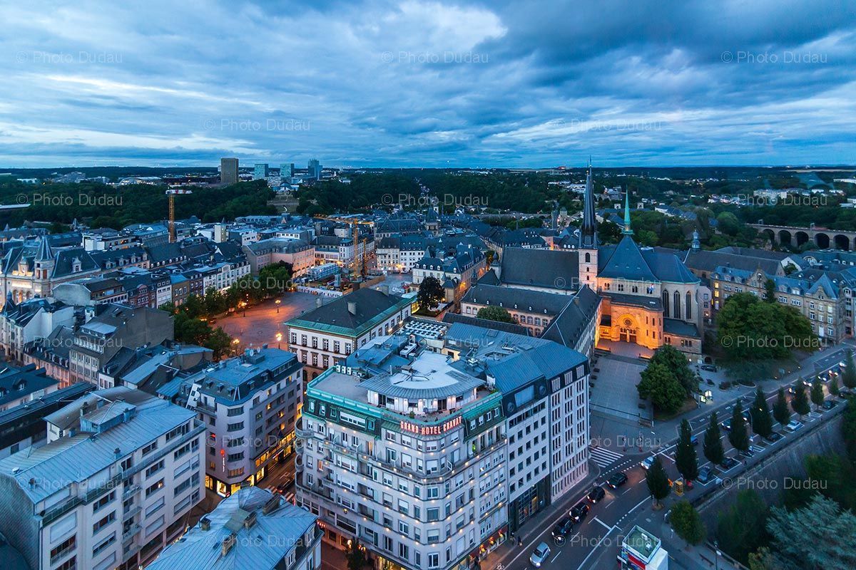 Aerial view of Luxembourg City at dusk, with illuminated buildings and a cloudy blue sky.