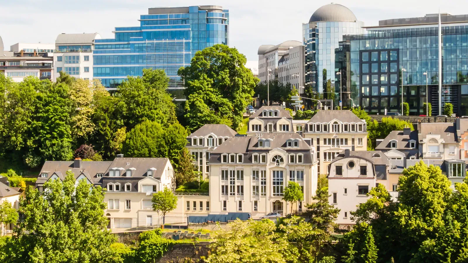 Luxembourg cityscape with modern and classic buildings, green trees, and a clear blue sky.