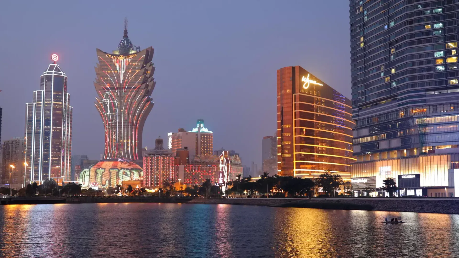 Night skyline of Macau, featuring illuminated casinos reflecting in the water. Buildings have varying shapes and colors, including gold and red.