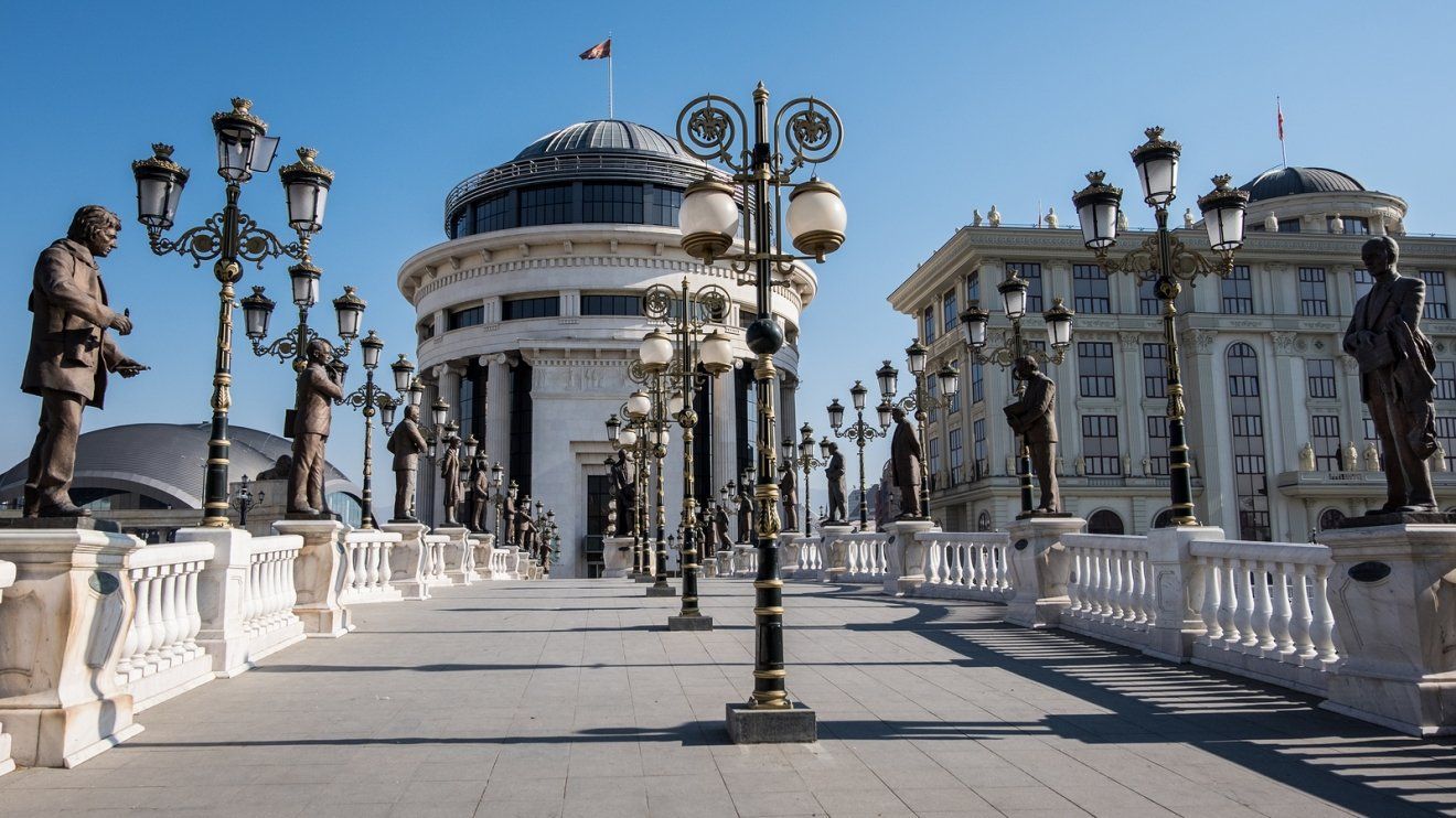 Bridge with ornate streetlights, statues, and neoclassical buildings against a clear blue sky in Skopje, North Macedonia.