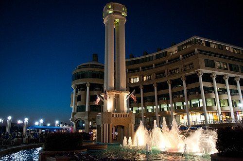 A nighttime view of a tall, lighted clock tower with a fountain and a building. Blue sky in the background.