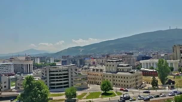 Cityscape of Skopje, North Macedonia with buildings, roads, and a mountain backdrop under a clear blue sky.