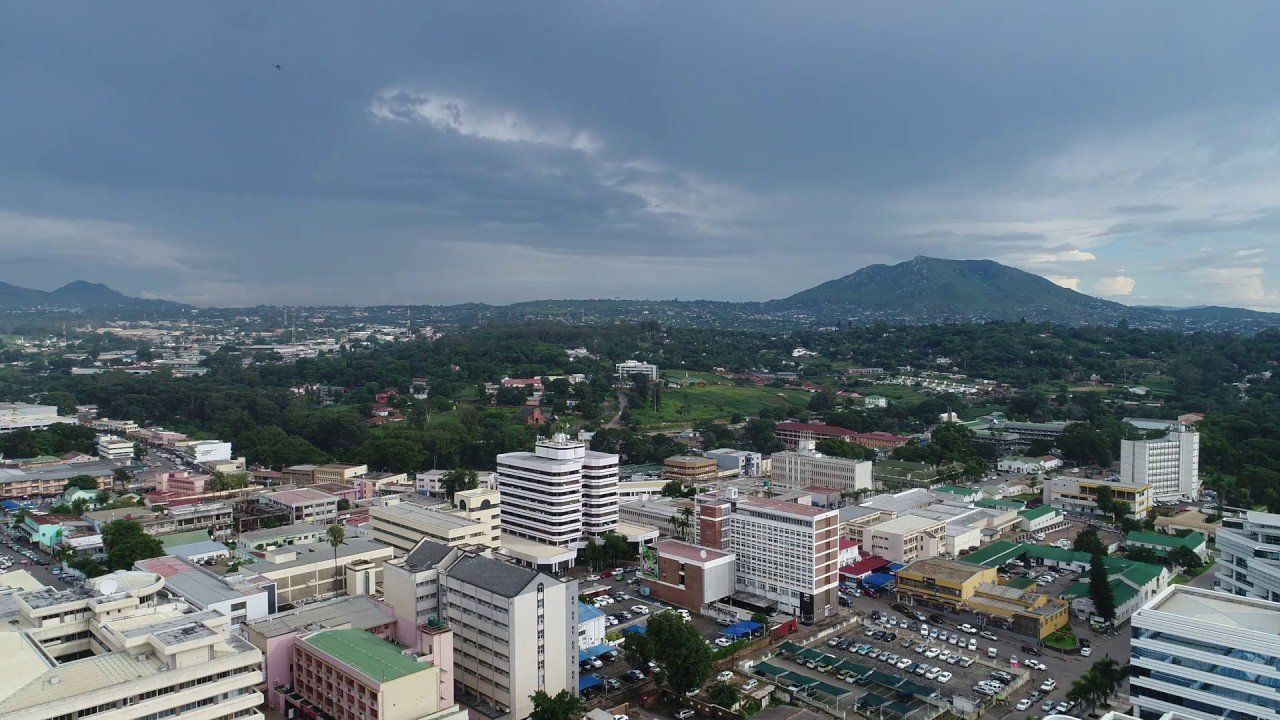 Overhead view of a city with white buildings and green trees, a mountain in the distance, and cloudy sky.