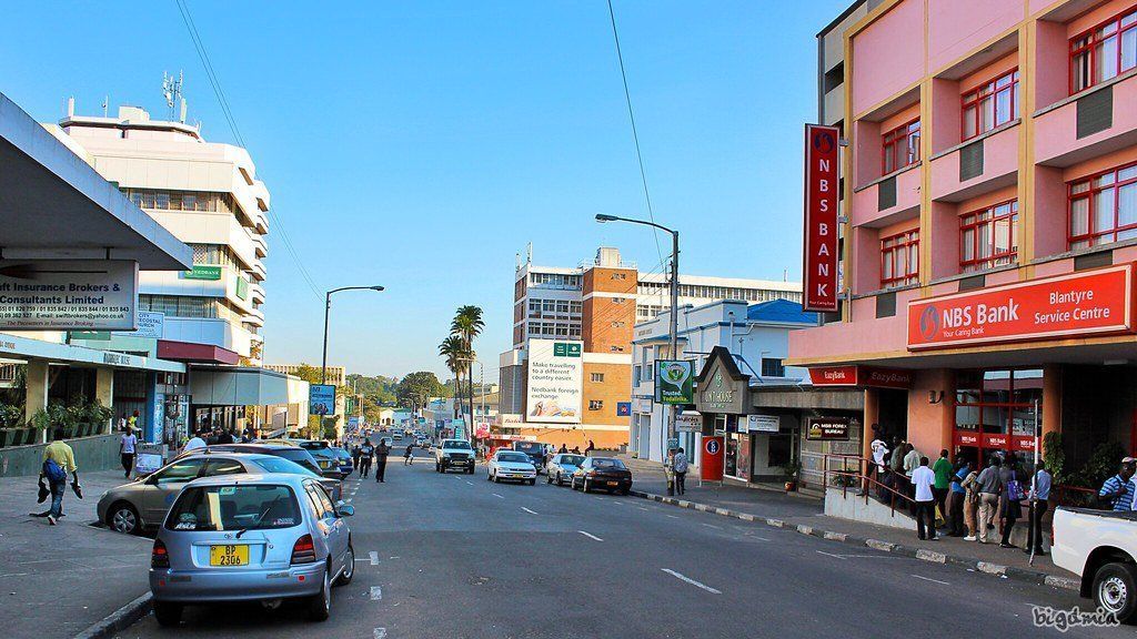 Street scene in a city with cars, pedestrians, and buildings including a hotel. Bright sky and sunny day.