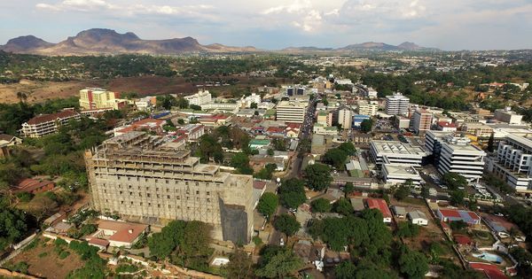 Aerial view of a city with a large, unfinished building in the foreground. Buildings and green spaces extend towards distant hills under a cloudy sky.