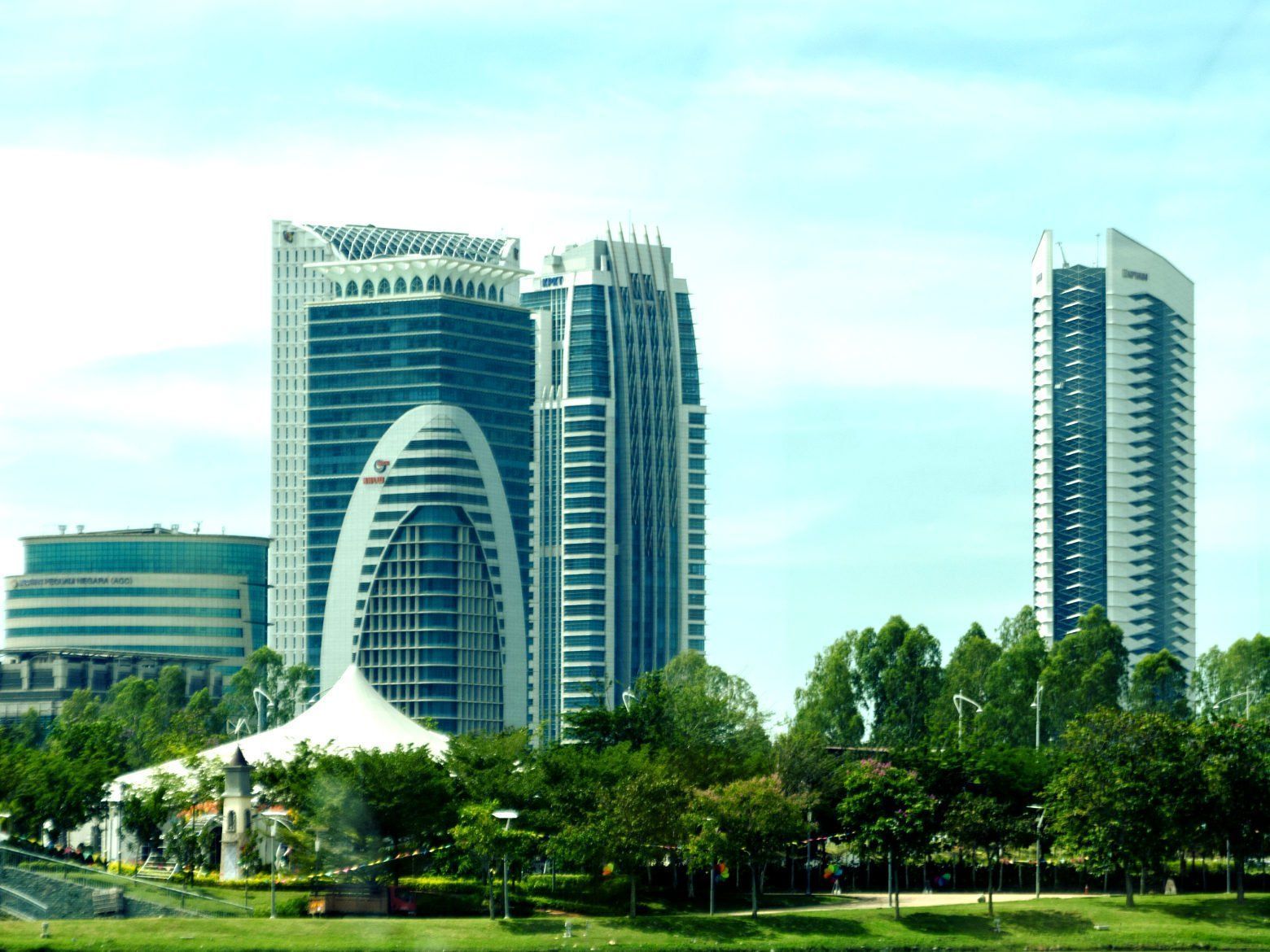 Modern skyscrapers against a bright sky, set amidst green trees and a white-roofed building in a park-like setting.