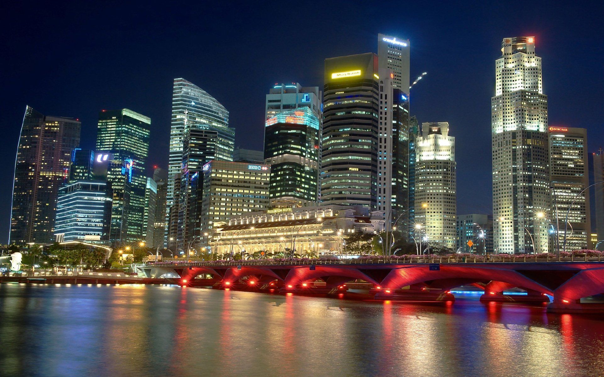 Night view of a cityscape in Singapore with brightly lit skyscrapers reflecting in the water below a red-lit bridge.
