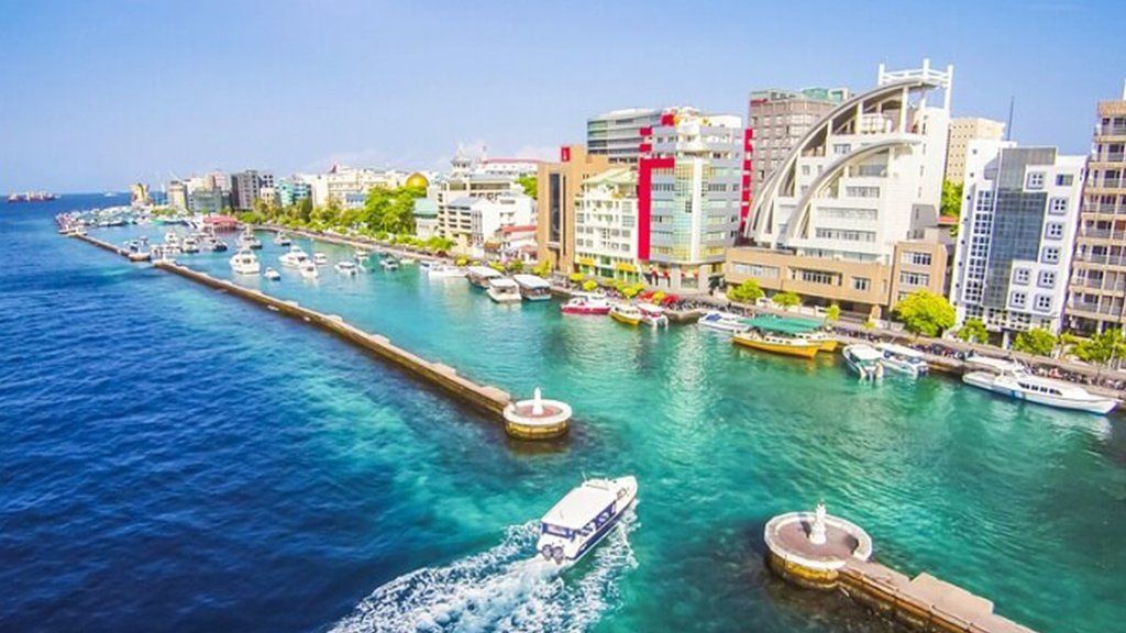 Aerial view of Male, Maldives, with buildings along the shore, boats in the turquoise water, and a sunny sky.