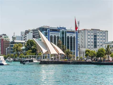 Waterfront view of Malé, Maldives, with modern buildings, boats, and a unique white-sail structure. Bright blue water under a sunny sky.