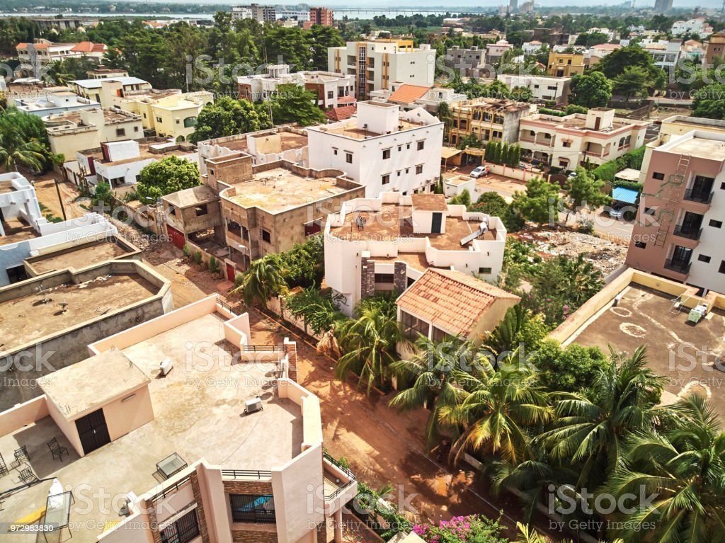 Aerial view of a sunny, densely-populated residential area with beige buildings, palm trees, and a body of water in the distance.