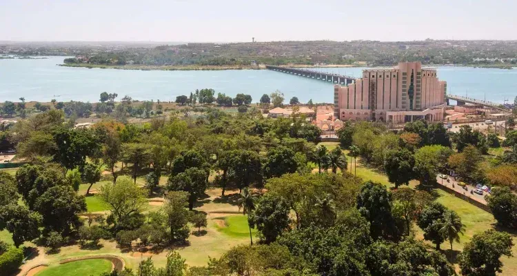 Overhead view of a city landscape with a large building and bridge over a body of water, surrounded by green trees and grass.