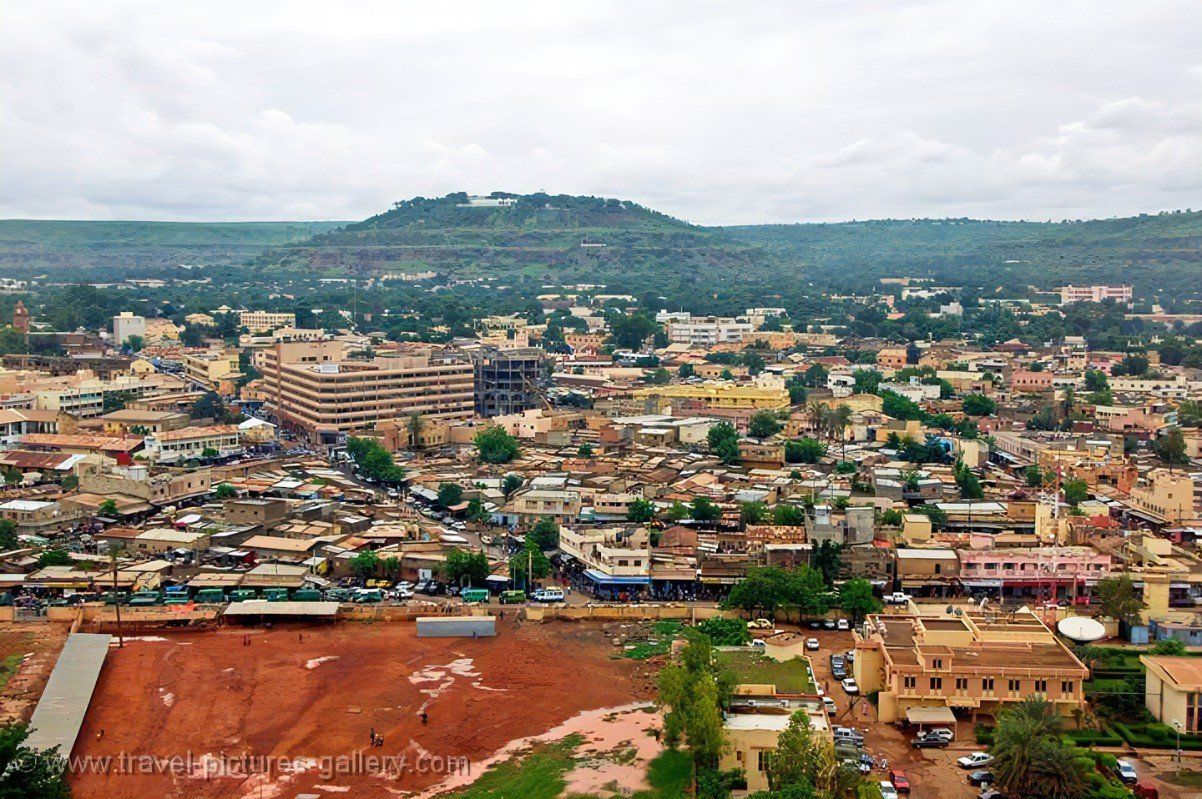 Aerial view of a cityscape with buildings, roads, and a large hill in the background. Overcast sky.