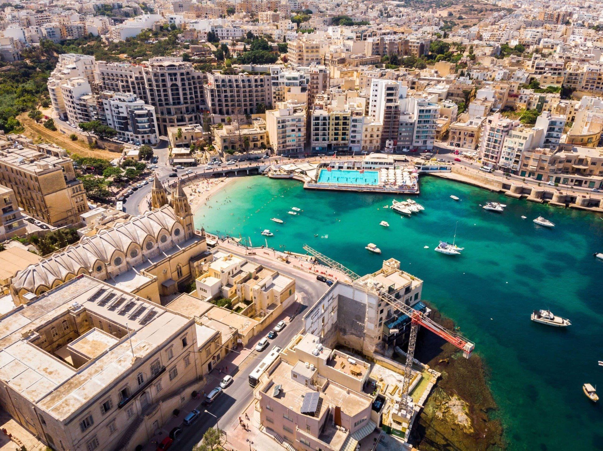 Aerial view of St. Julian's Bay, Malta, showing a turquoise bay surrounded by buildings, boats, and a swimming pool.