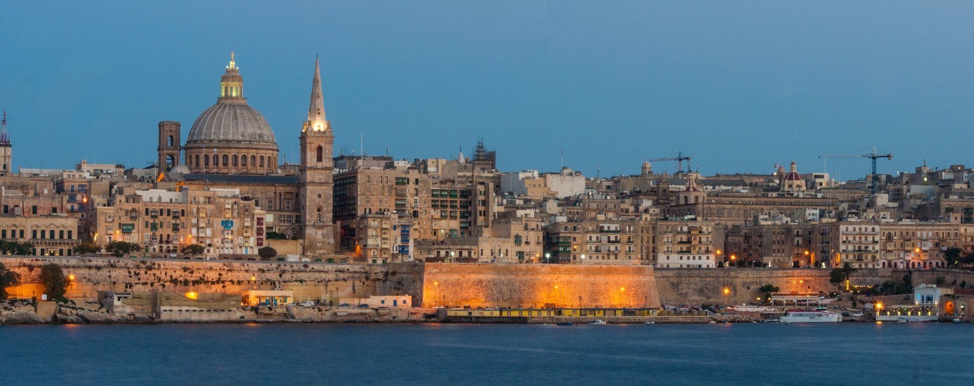 City skyline at dusk with a large domed church and tall steeple, illuminated buildings line the waterfront, and a calm blue sea.