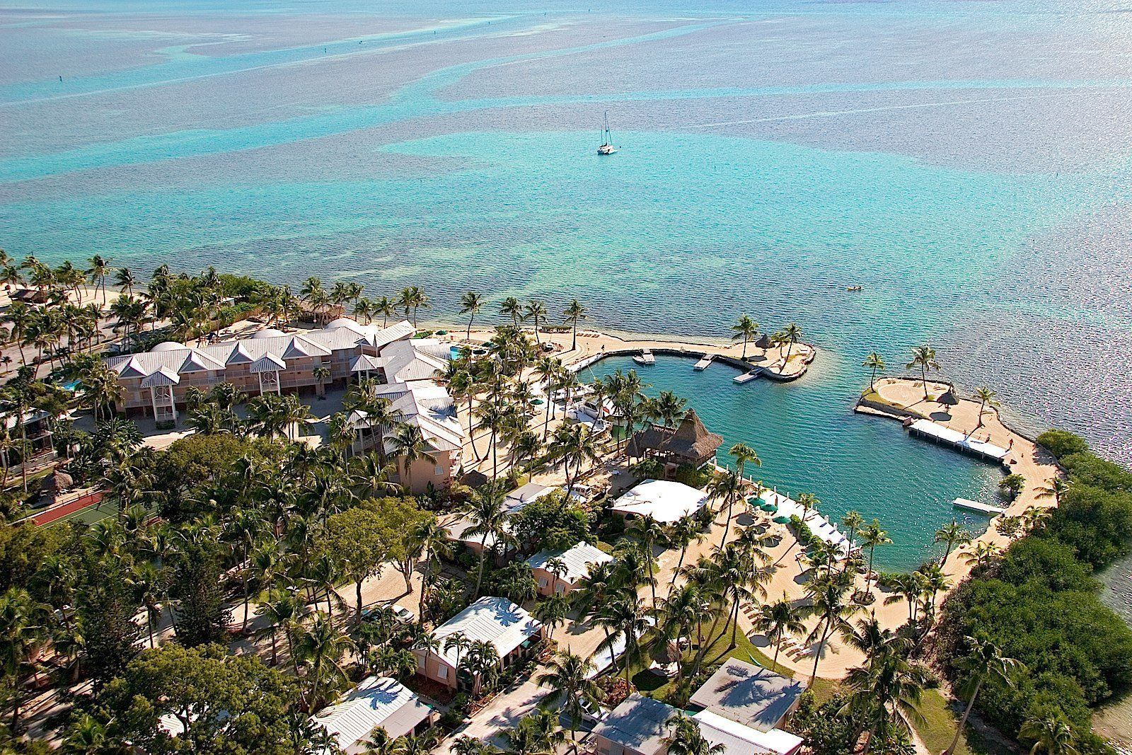 Aerial view of a resort on a tropical island. Turquoise water surrounds a complex of buildings and palm trees, with a small harbor.