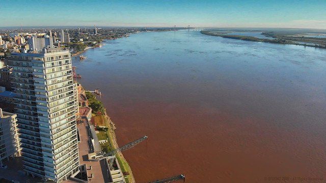 High-angle view of a cityscape bordering a wide, brown river. A tall apartment building is on the left, and the river stretches into the distance.