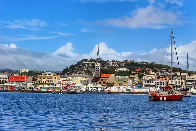 Waterfront cityscape with a church spire on a hill. A red sailboat is in the foreground, with colorful buildings and blue sky.