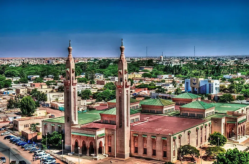 Mosque with two tall minarets in a city setting under a clear blue sky. Buildings are primarily brown with green rooftops.