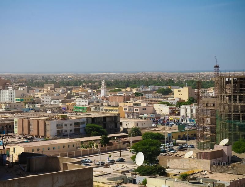 Cityscape of Nouakchott, Mauritania, with low-rise buildings in shades of tan and brown under a blue sky. A white minaret is visible.