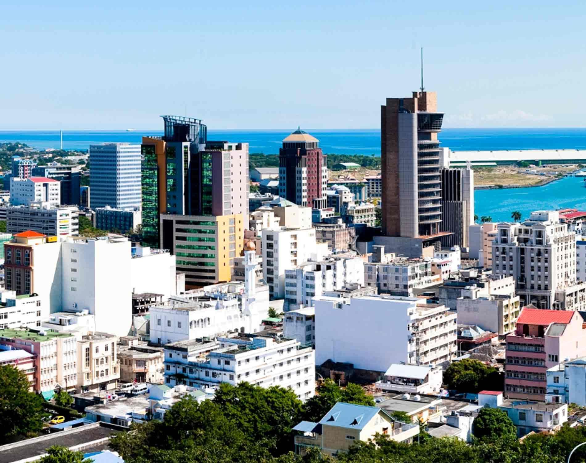 City skyline under a bright blue sky, featuring modern high-rise buildings and the ocean in the background.