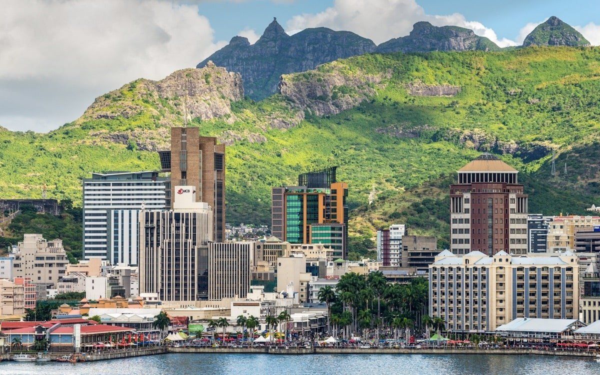 Cityscape in Mauritius with buildings of various heights along a waterfront, backed by lush green mountains under a blue sky.