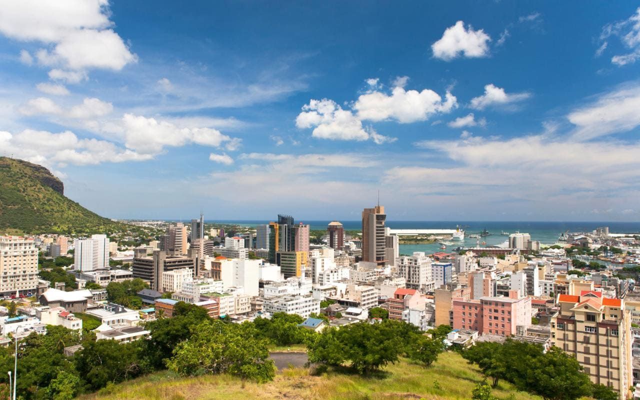 City skyline under a blue sky with fluffy clouds, buildings of varying heights, lush green hills in the foreground and the ocean in the distance.