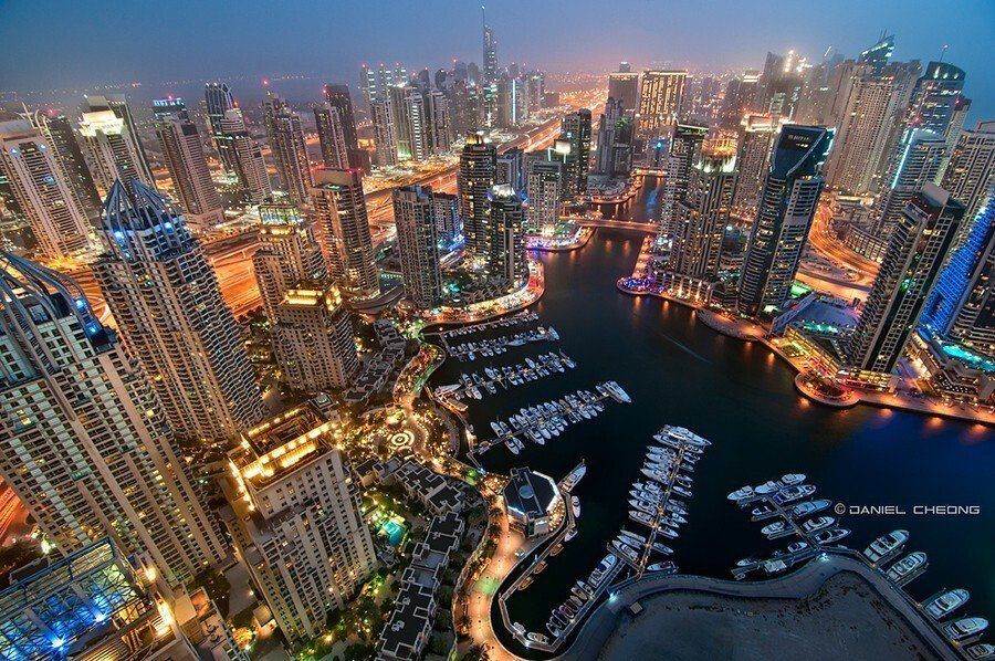 Aerial view of Dubai Marina at night, featuring numerous illuminated skyscrapers, a boat-filled harbor, and city lights.