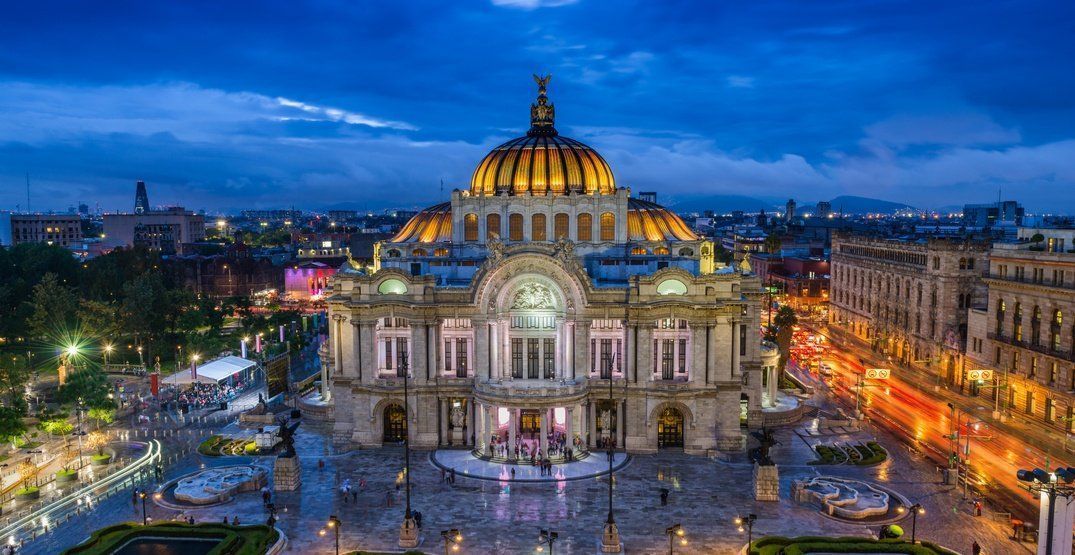 Palacio de Bellas Artes in Mexico City at dusk, illuminated against a blue sky. People gather in the plaza below.