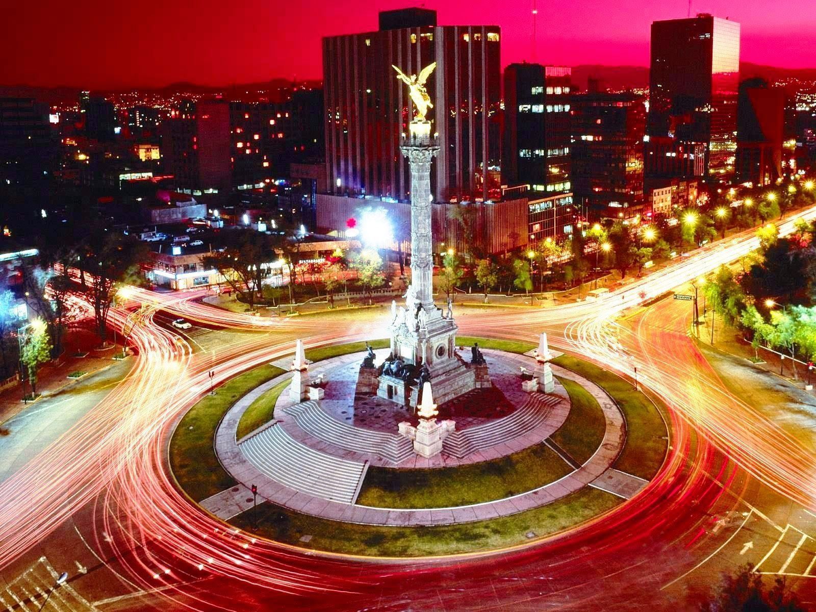 Nighttime view of the Angel of Independence monument in Mexico City, with blurred streaks of car lights around the roundabout. Buildings and illuminated lights glow in the background.