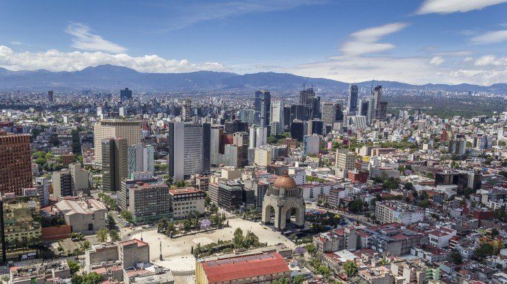 Aerial view of Mexico City with a mix of buildings, including skyscrapers and historic architecture, under a blue sky. Mountains are visible in the distance.