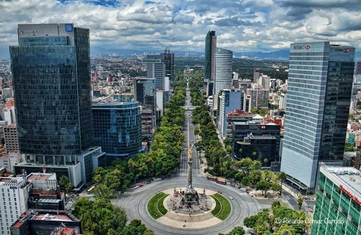 Aerial view of Mexico City with skyscrapers, a tree-lined boulevard, and a monument in a roundabout under a cloudy sky.