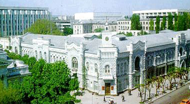 Ornate, gray-and-white classical building with a clock tower, likely a university, in an urban setting, with greenery and other buildings.