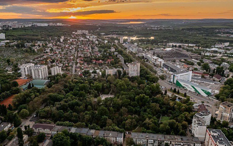 Aerial view of a city at sunset. Buildings and trees dominate the scene, with an orange and yellow sky in the background.